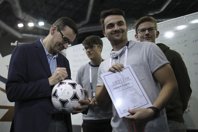Premier Mateusz Morawiecki wśród zwycięzców hackathonu w Krakowie podpisuje piłkę nożną.