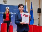 Minister Izabela Leszczyna prezentuje konkursowy dyplom (w tle rzędy czerwonych, papierowych torebek z upominkami dla laureatów)