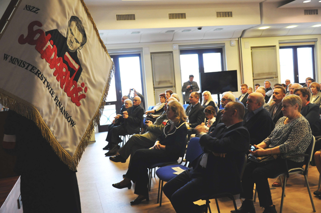 Uczestnicy konferencji słuchają prelegentów, po lewej stronie sztandar Solidarności z wizerunkiem księdza Popiełuszki. 