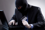 Zdjęcie mężczyzny w kominiarce przed komputerem z kartą kredytową w ręce