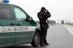 Na zdjęciu funkcjonariusz Służby Celno-Skarbowej patrzący przez lornetkę, z lewej strony widoczny samochód patrolowy